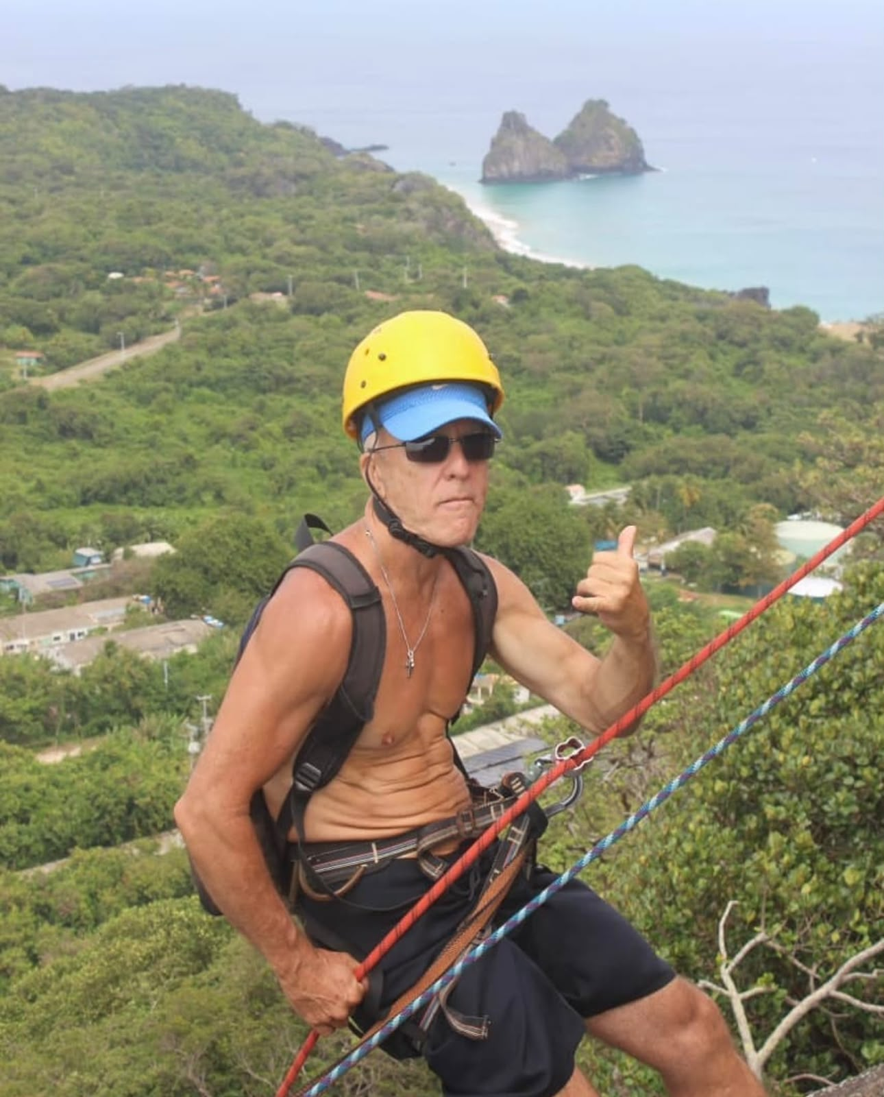 Alpinista com capacete amarelo sinalizando durante o rapel, com a praia ao fundo